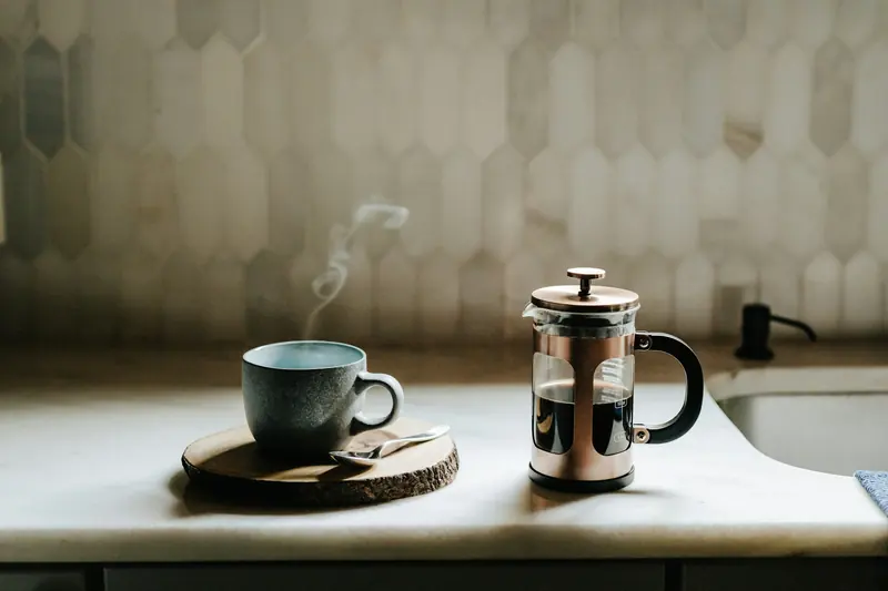 Steaming ceramic cup on a wood slice beside a steel French press on a pale stone kitchen counter, hexagonal tile backsplash, soft daylight.